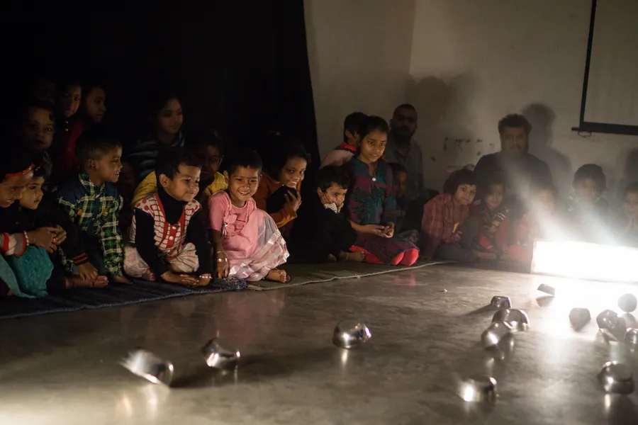 Rows of small children sitting close together on the floor in a darkened room, watching.