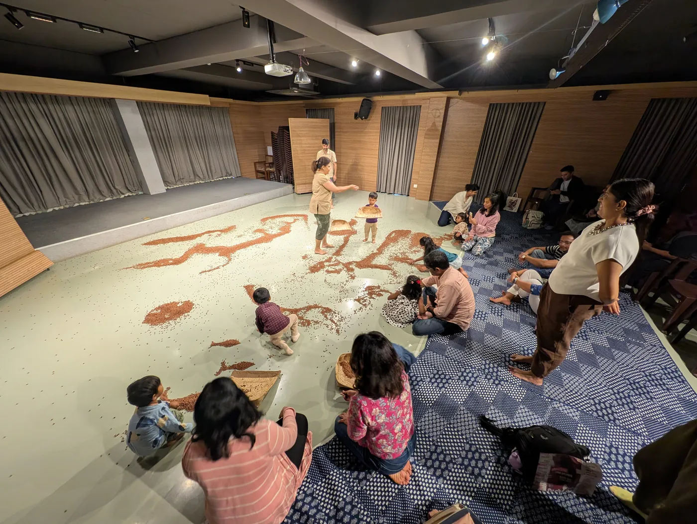 After the play, children and adults kneel together on the floor amid the spread of black gram, handling it.