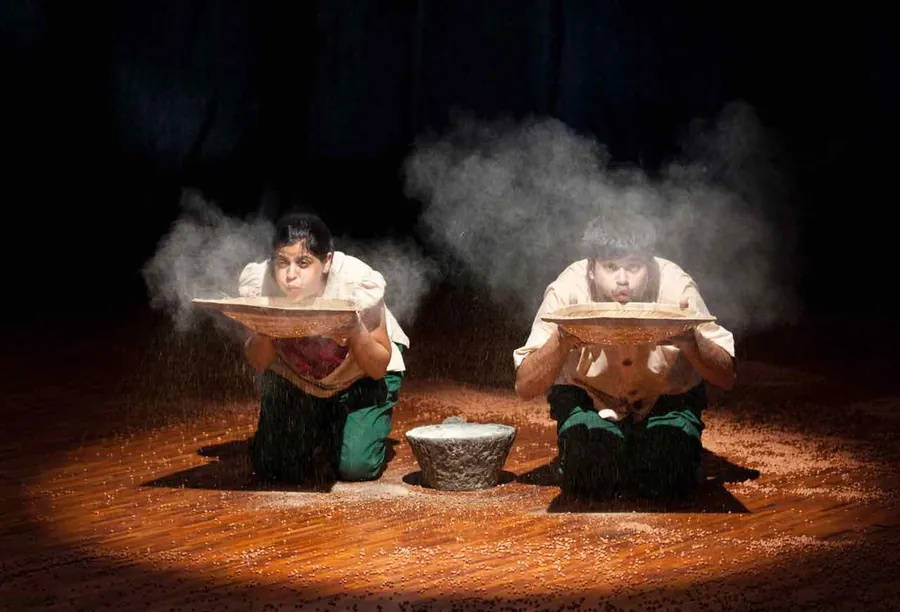 Two performers kneel in darkness as black gram falls through a shaft of light, throwing up a cloud of dust.