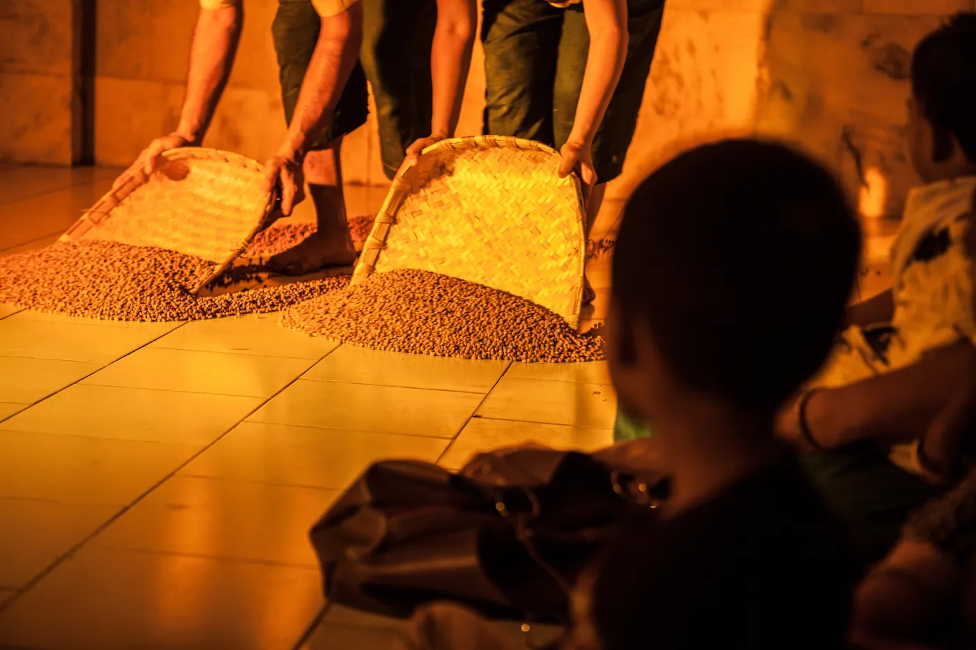 Black gram heaped and spread across a floor in warm low light, a child's silhouette at the edge of the frame.