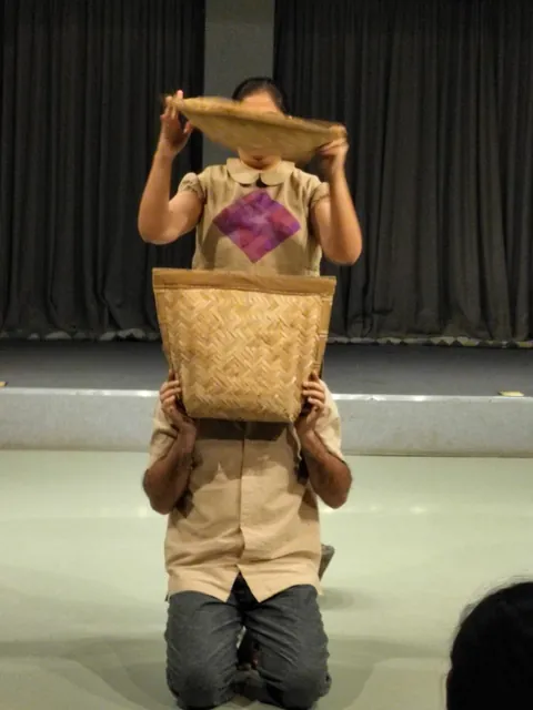 A performer kneels holding a woven winnowing basket up to her face.