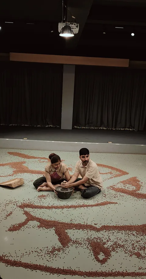 Two performers sit together inside a wide spiral of black gram laid across the floor.