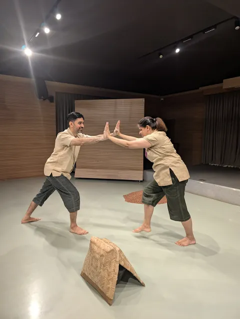 Two performers lean towards each other, palms meeting, a woven basket on the floor between them.