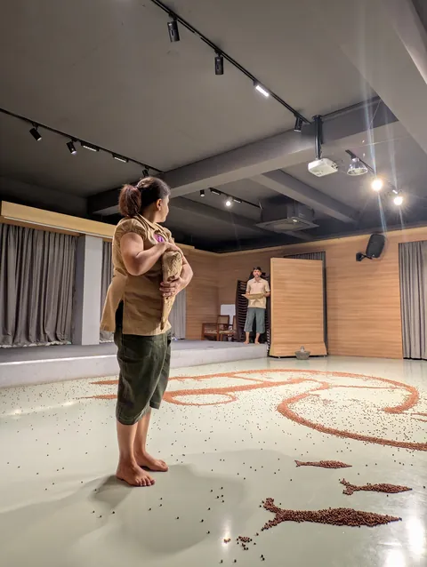 A performer stands at the edge of the spread of gram, looking down at it.