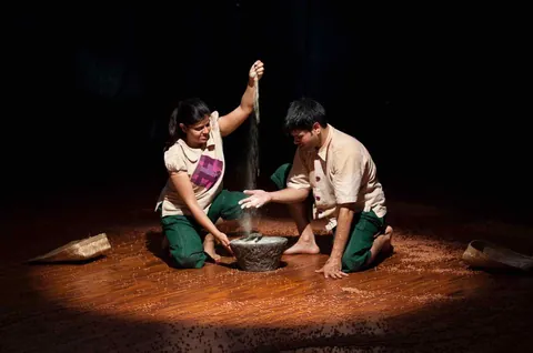 Two performers kneel in a pool of light, one pouring gram into a stone bowl.