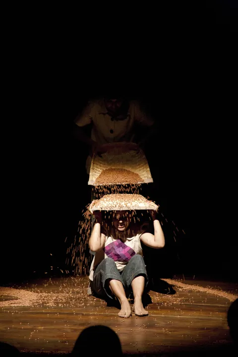 A performer tips a basket of gram over her head, the grain falling through the light.