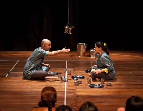 Two performers kneel among steel vessels on the wooden stage.