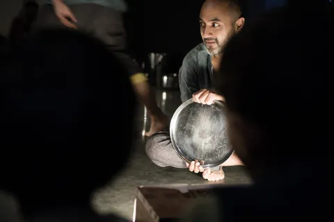 A performer kneels beside a large steel vessel in the dark.