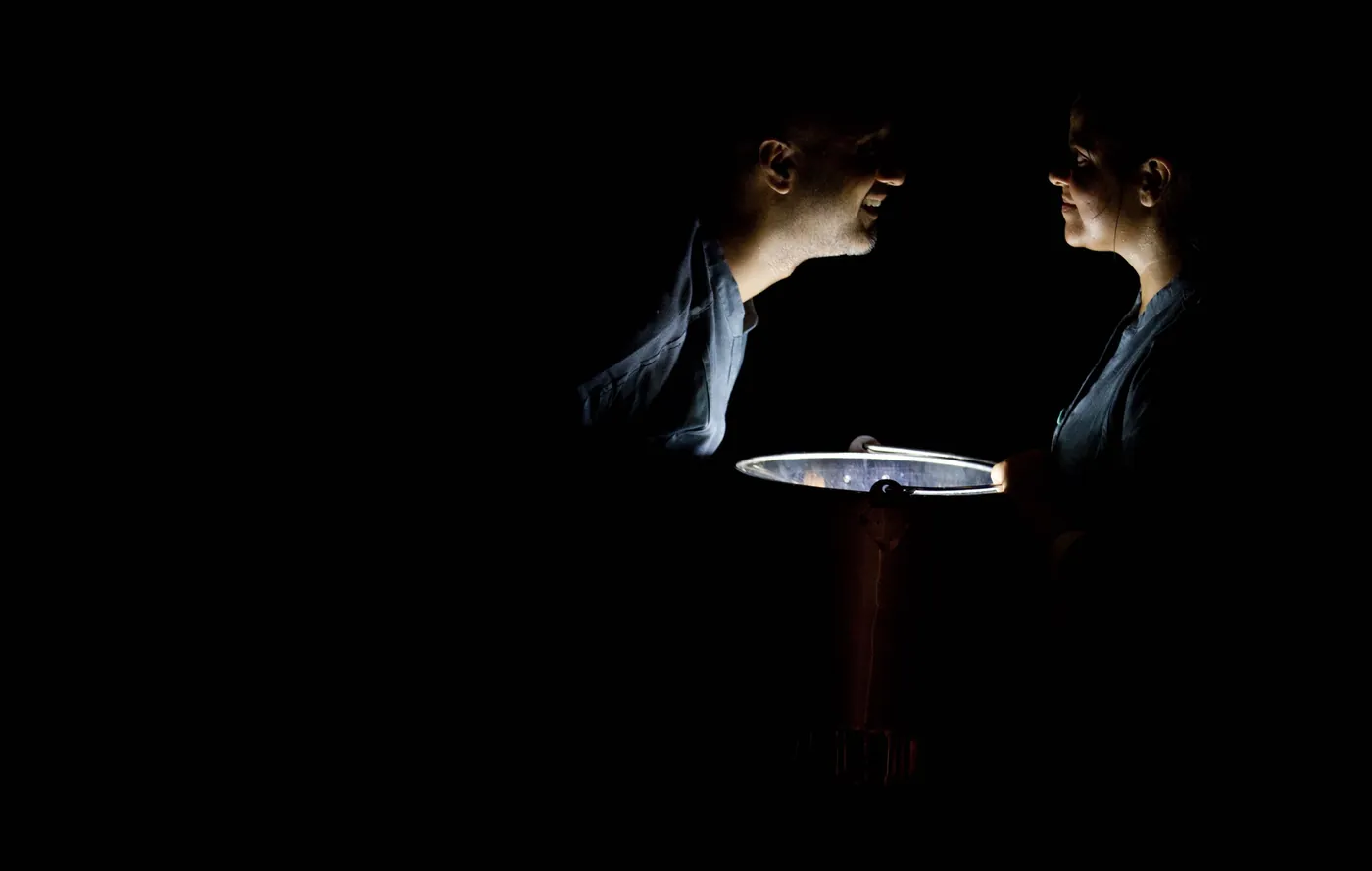 In near darkness, two performers hold a large steel plate between them, lit from below.