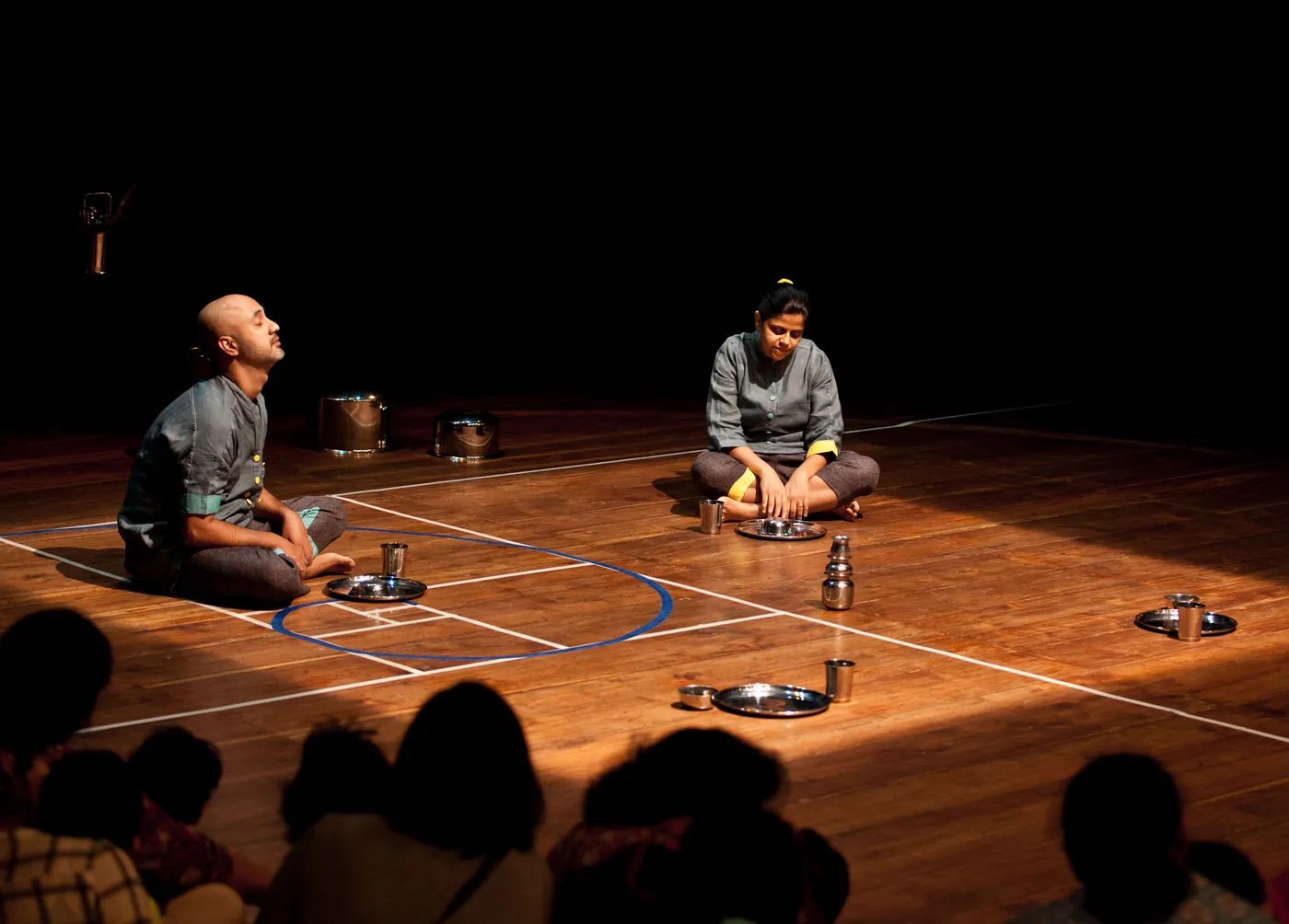 Wide view of the stage: two performers kneel on a wooden floor among steel vessels, the audience silhouetted in the foreground.