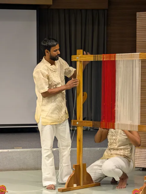 A performer stands at a tall wooden loom threaded in red and white while another crouches at its base.