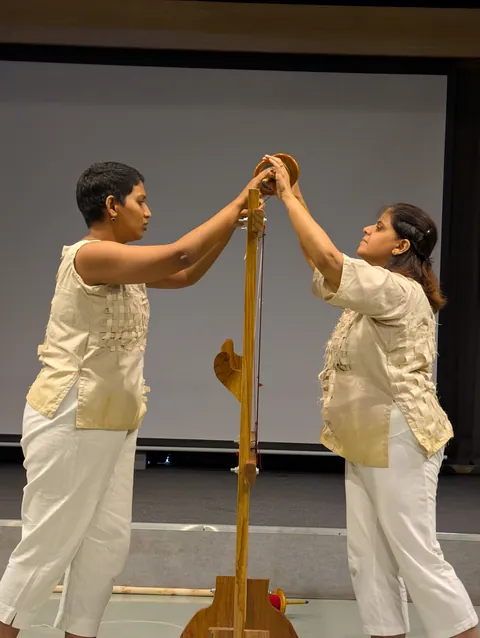 Two performers face each other across an upright wooden post, their hands meeting at the top.