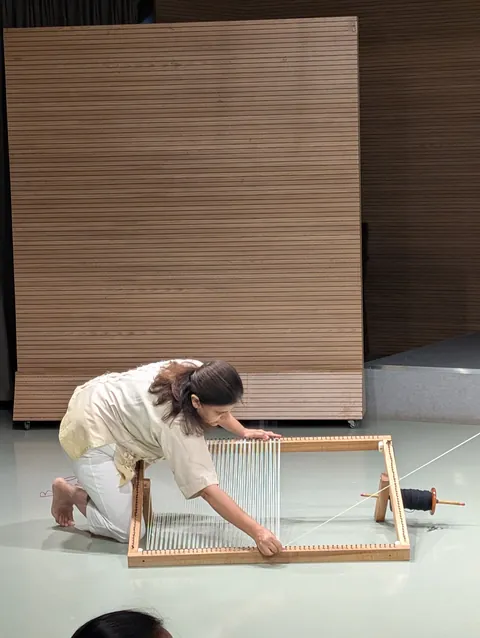 A performer kneels on the floor beside a small frame loom, drawing thread across it.