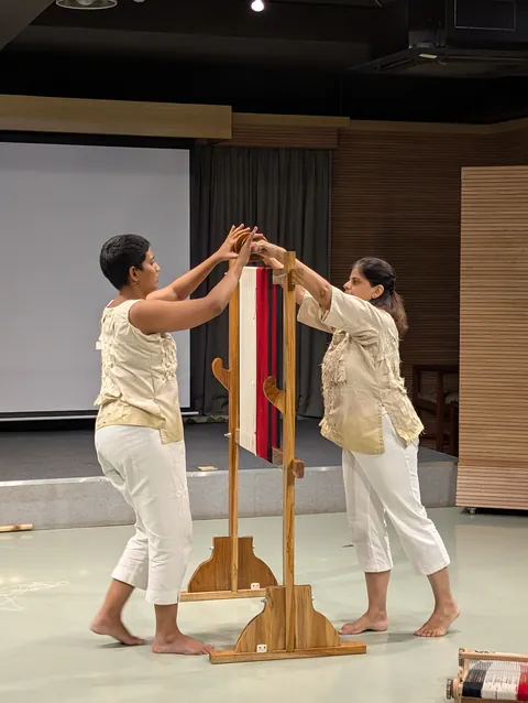 Two performers reach up together at the tall loom, its red and white warp between them.