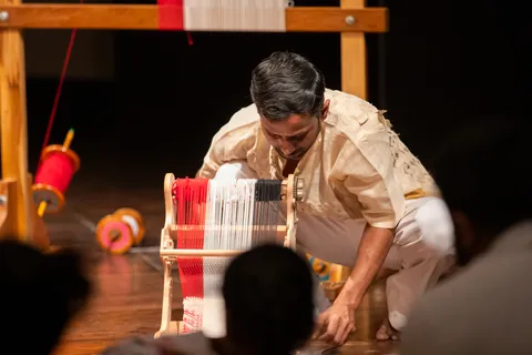 A performer crouches over a low frame loom, the audience silhouetted in the foreground.