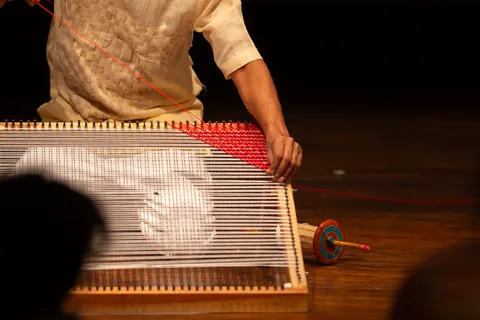 Close on a performer&rsquo;s hands laying red thread across the white warp of a low loom.
