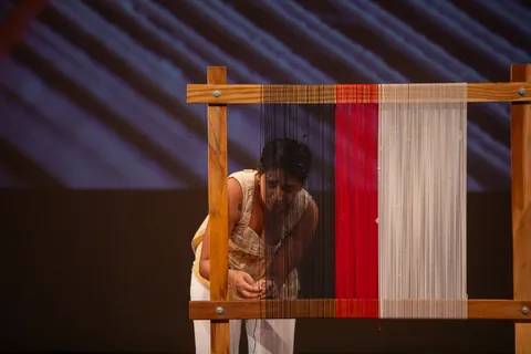 A performer stands behind the tall loom, seen through its red and white threads.