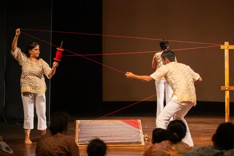 A performer holds a red spool aloft as long threads radiate across the stage.