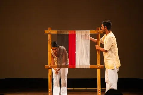 Two performers work at either side of the tall loom, one bending low.