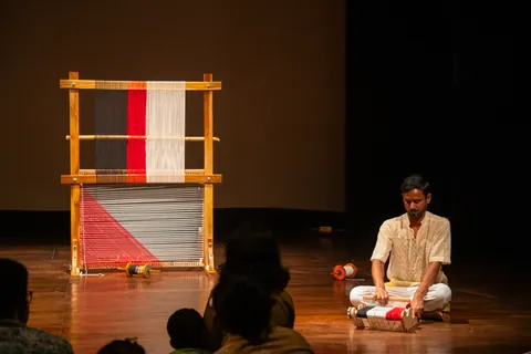A performer sits on the floor beside the finished weave, the audience watching from the dark.