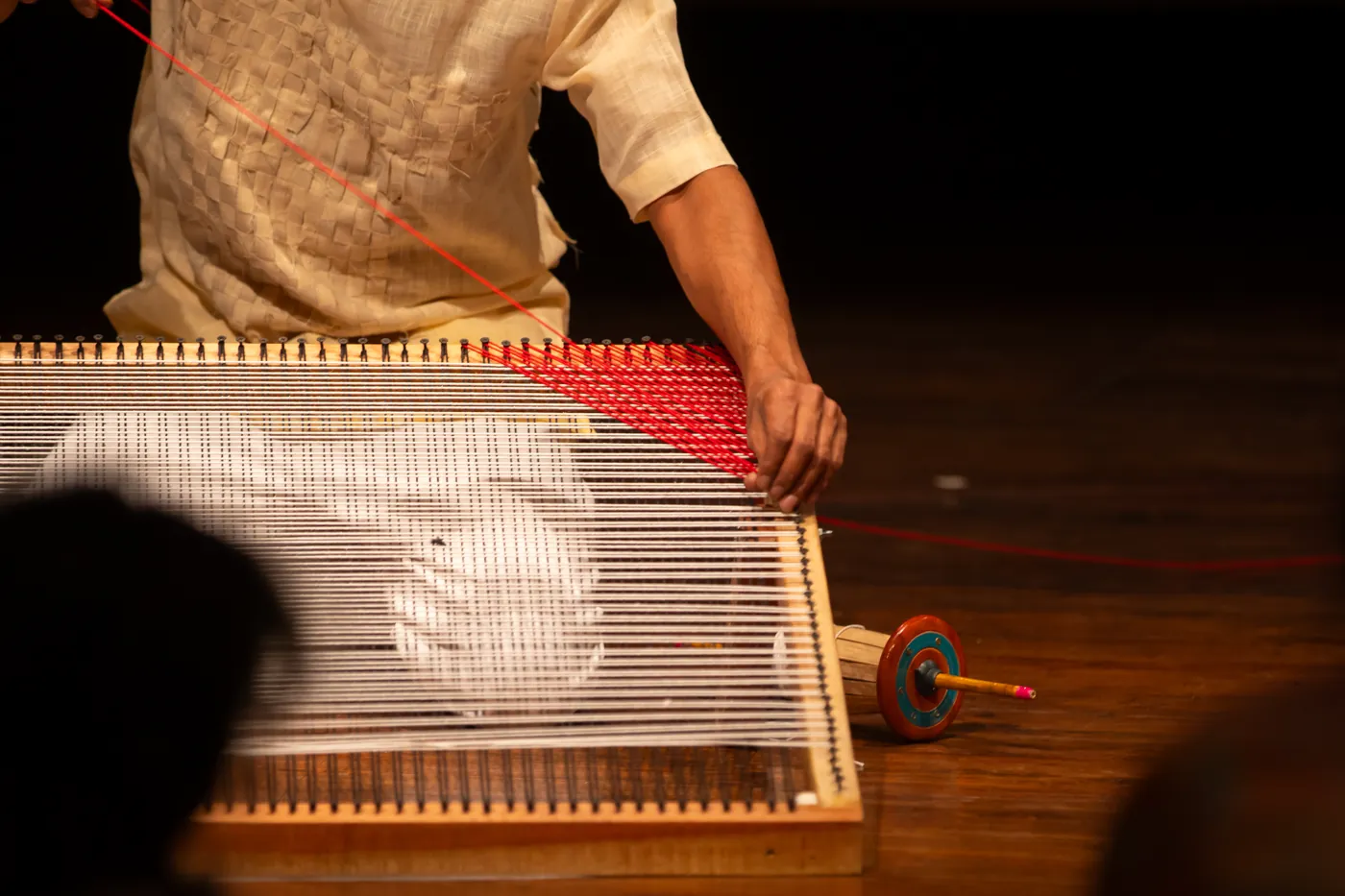 A performer's hands working red and white thread across the frame of a low wooden loom.