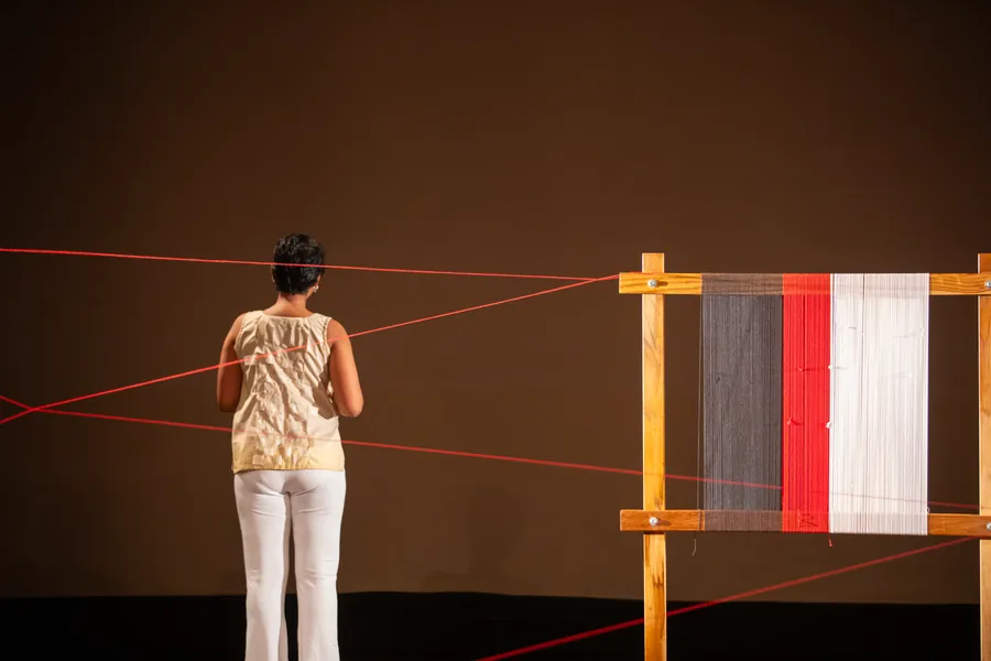 Red threads stretched in long radiating lines across a dark stage between a performer and a wooden loom.