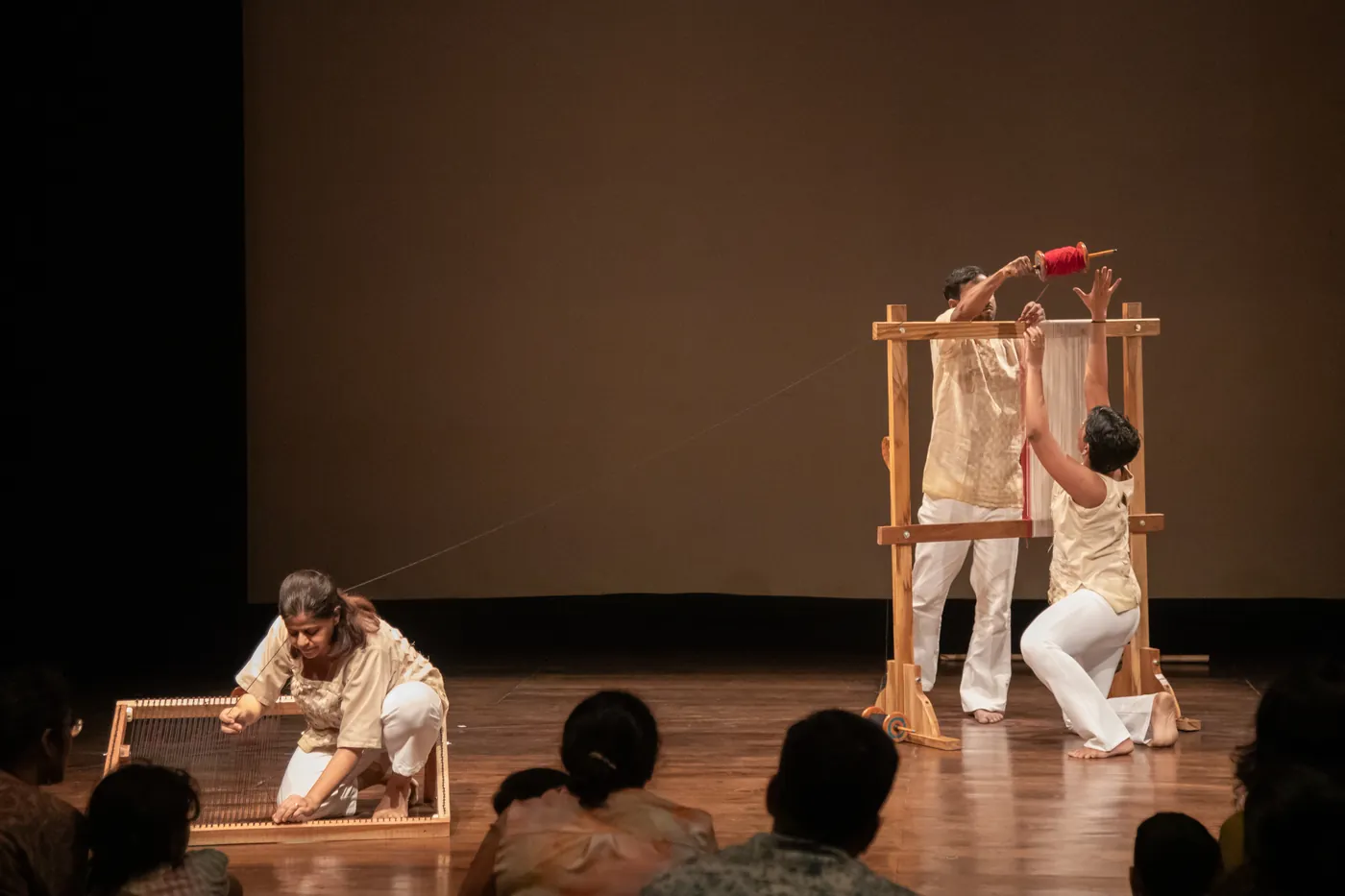 Wide view of the stage with three performers at separate looms, the audience silhouetted along the front edge.